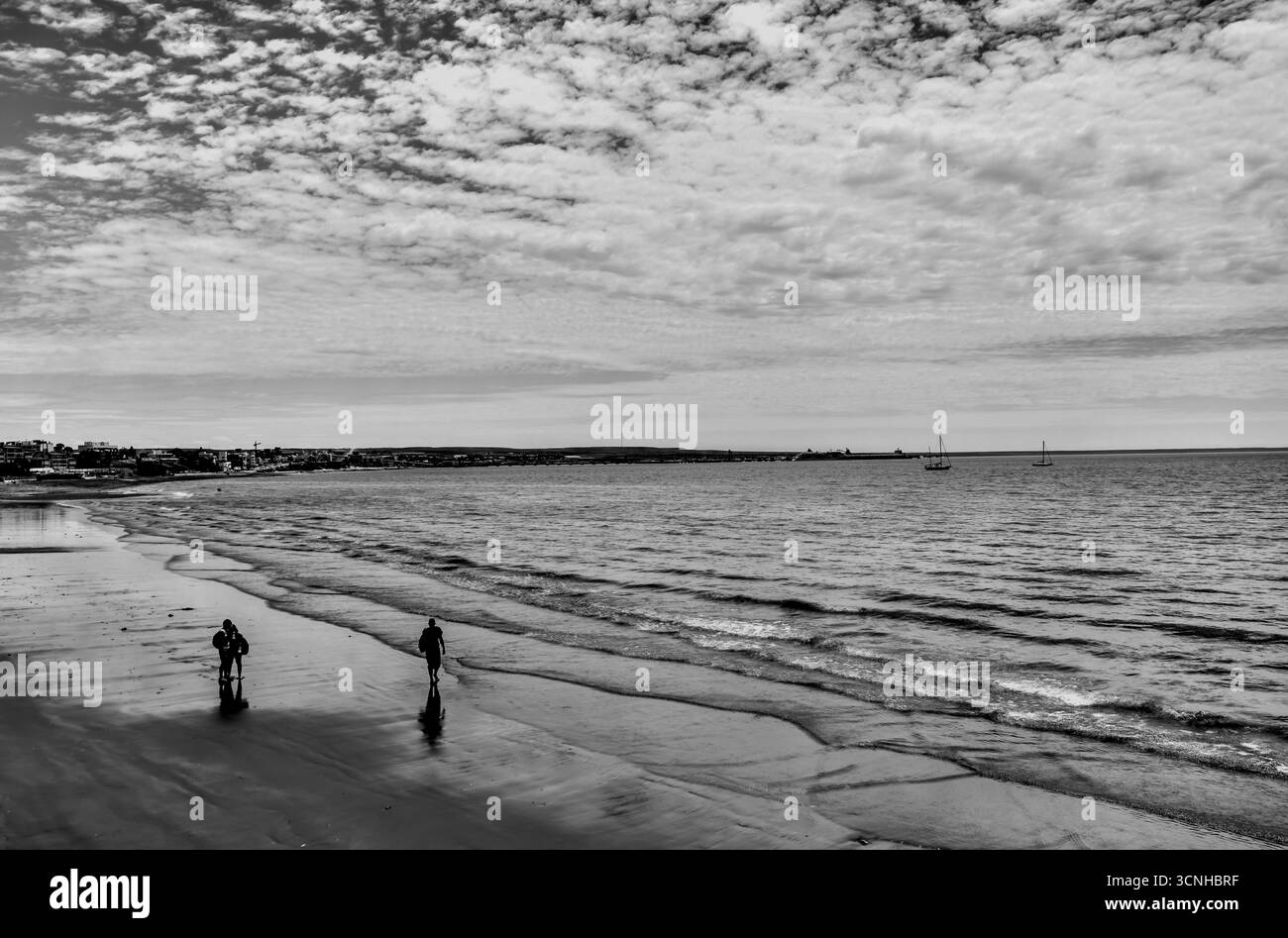 Two figures walk along the vast, windswept beach of Puerto Madryn as moody clouds stretch over the Patagonian shoreline. Stock Photo