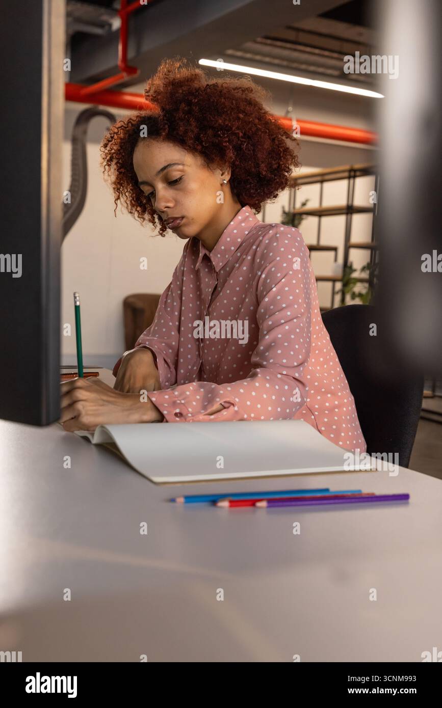 African american woman writing in notebook at office desk with computer monitor and colored pencils Stock Photo