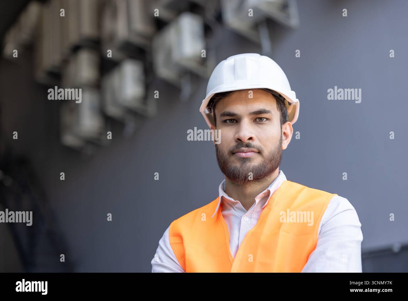 Young bearded man standing outdoors, wearing a white hard hat and an orange safety vest, representing construction, engineering, safety, and a profess Stock Photo
