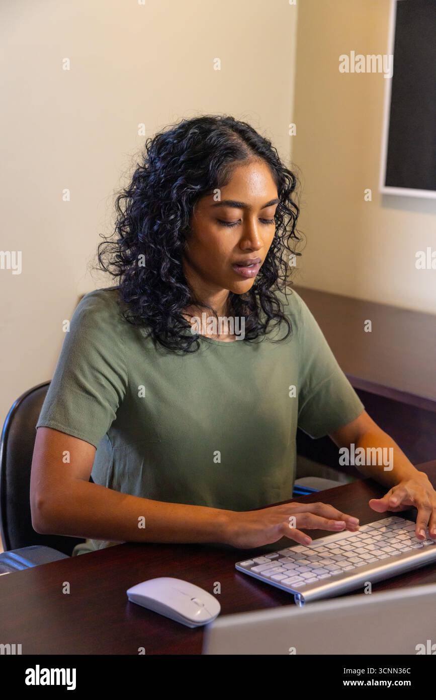Indian woman typing on keyboard at office desk with wireless mouse focusing on monitor Stock Photo