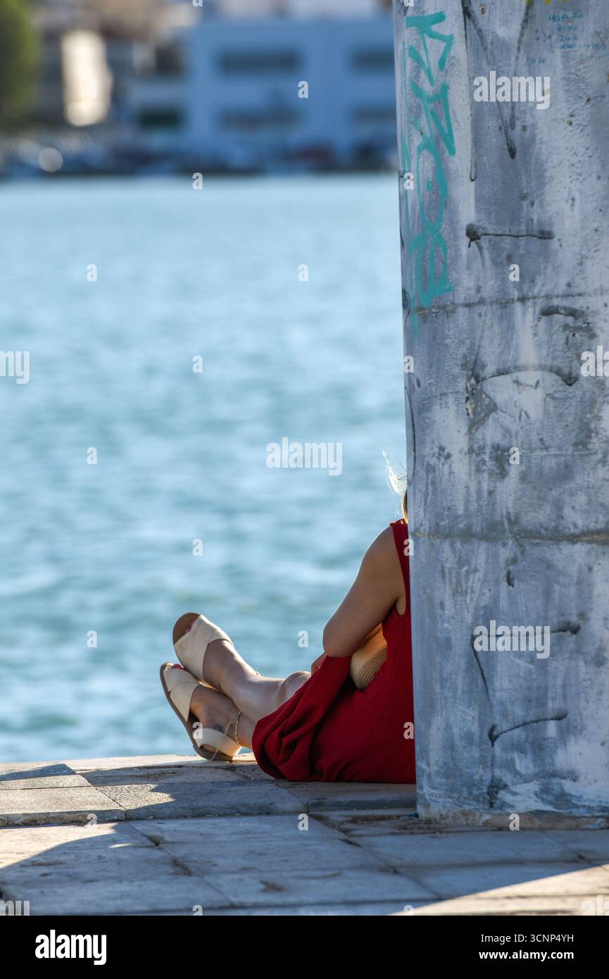 young woman sitting alone wearing a red dress leaning on a wall looking out to sea on a warm mediterranean evening in Zante town, Zakynthos, Greece Stock Photo