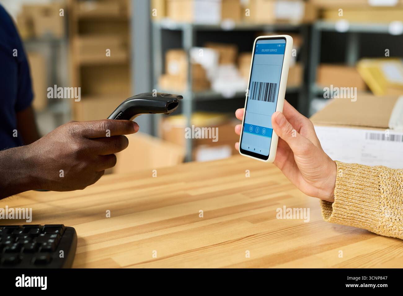 Caucasian young adult woman holding smartphone displaying barcode while Black man using handheld barcode scanner, both interacting at wooden desk in warehouse or shipping facility Stock Photo