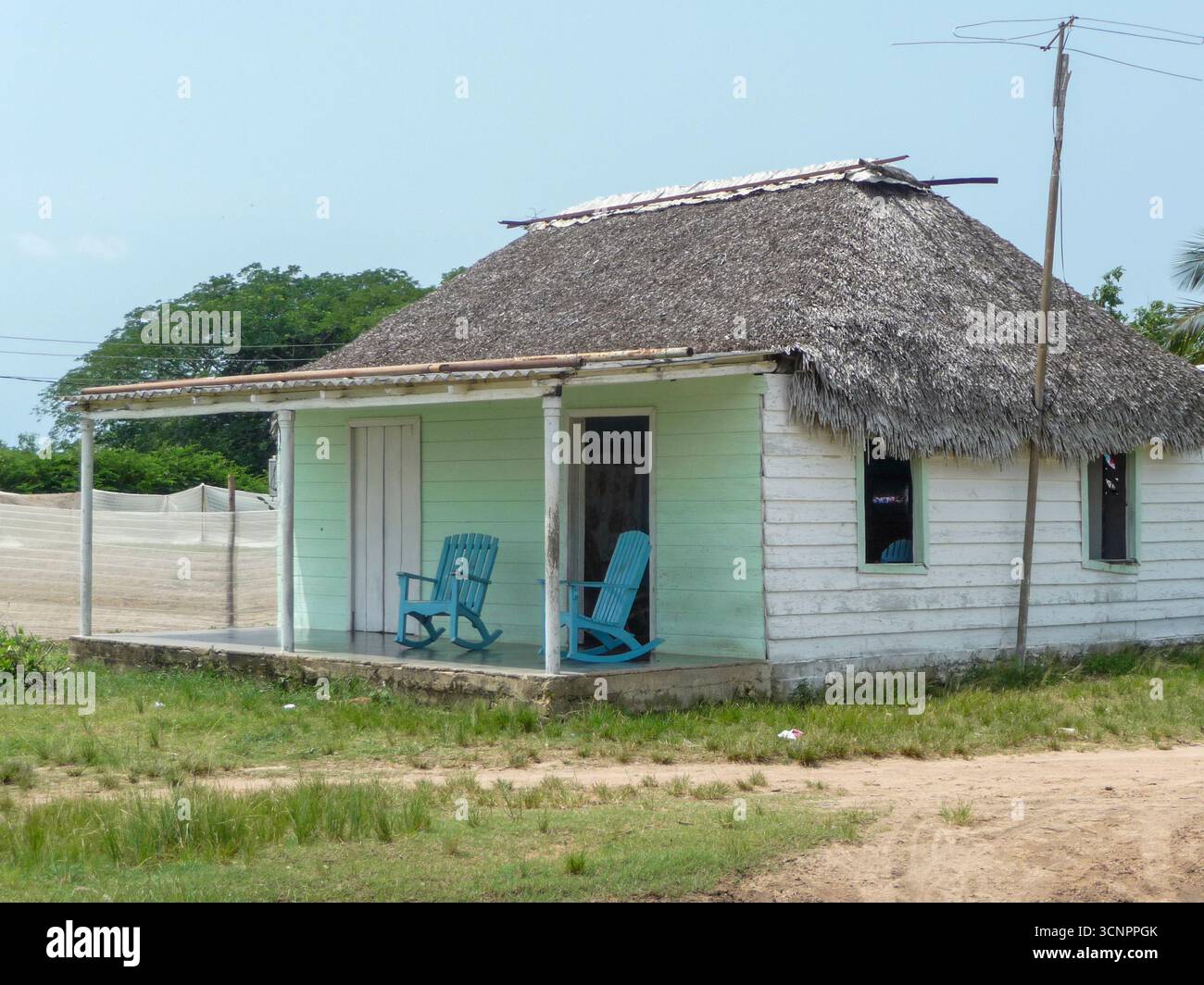 Cuba - Rural house - Weathered boards and thatch shelter blue rocking chairs in a sunlit clearing Stock Photo