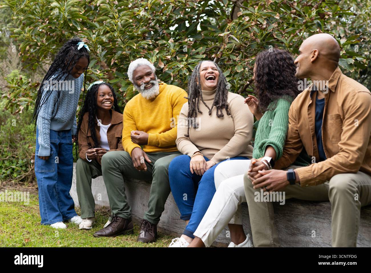 Diverse family gathering on wooden log bench in park wearing autumn sweaters sporting smartwatch Stock Photo
