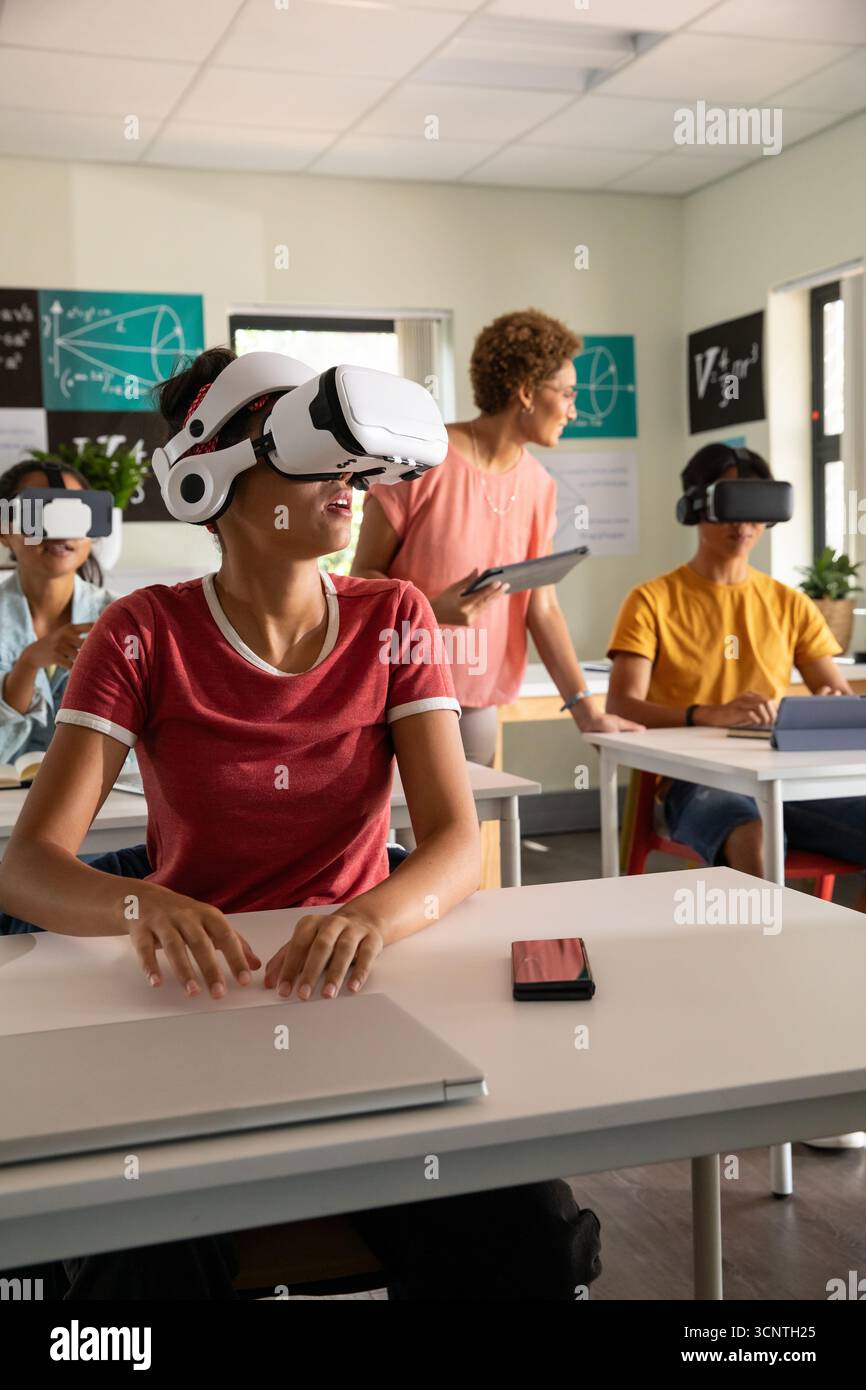 Diverse classmates wearing vr headsets using laptops while teacher with tablet in classroom Stock Photo