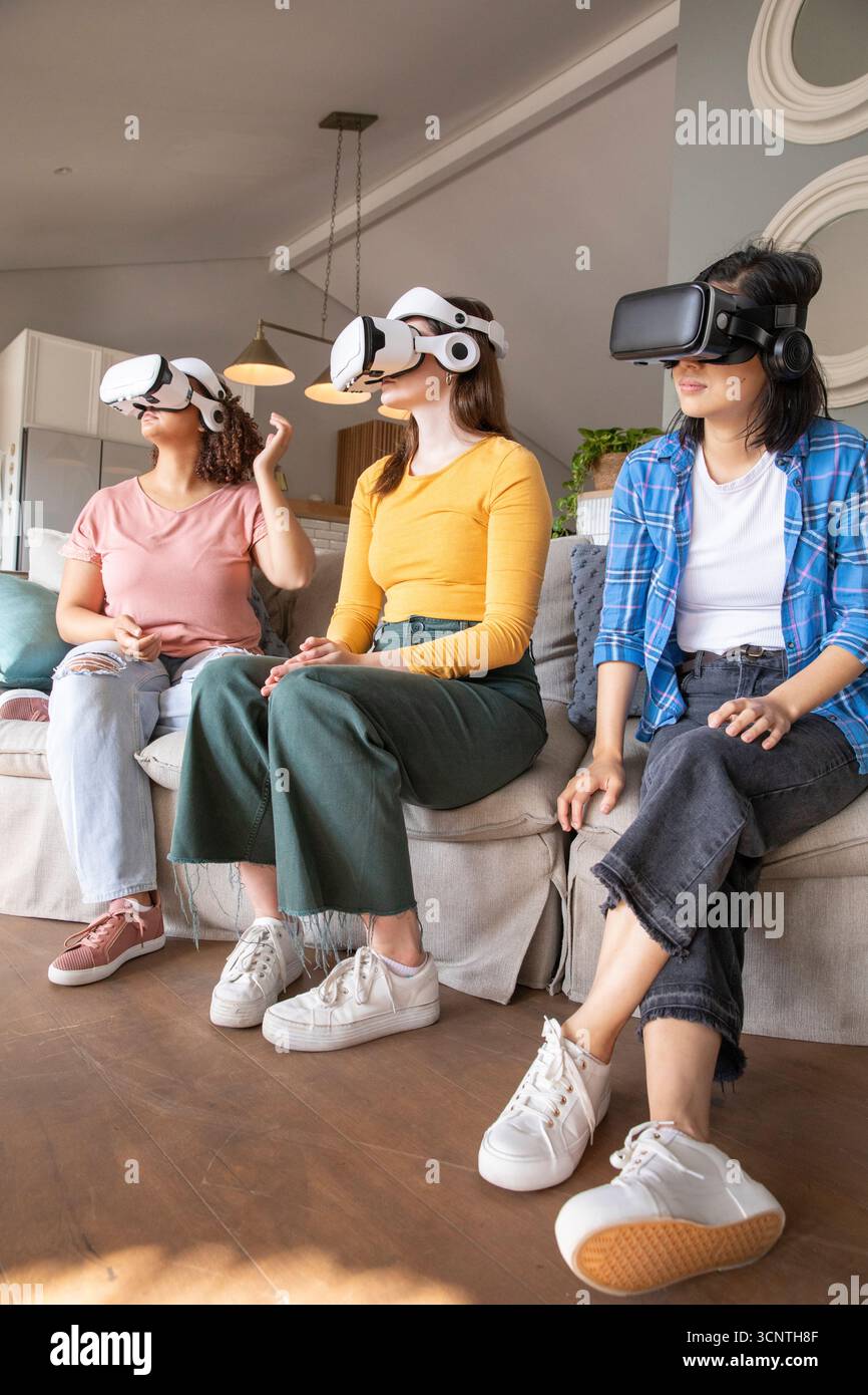 Diverse female friends sitting on sofa engaging with virtual reality headsets in modern living room Stock Photo