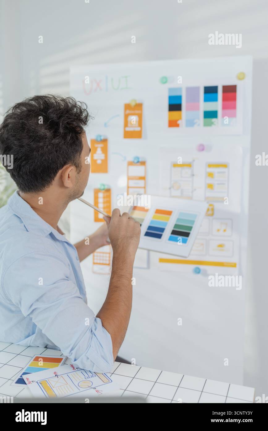A focused designer is working on UX/UI design ideas, analyzing color palettes and interface structures pinned on a board. The scene reflects creativit Stock Photo