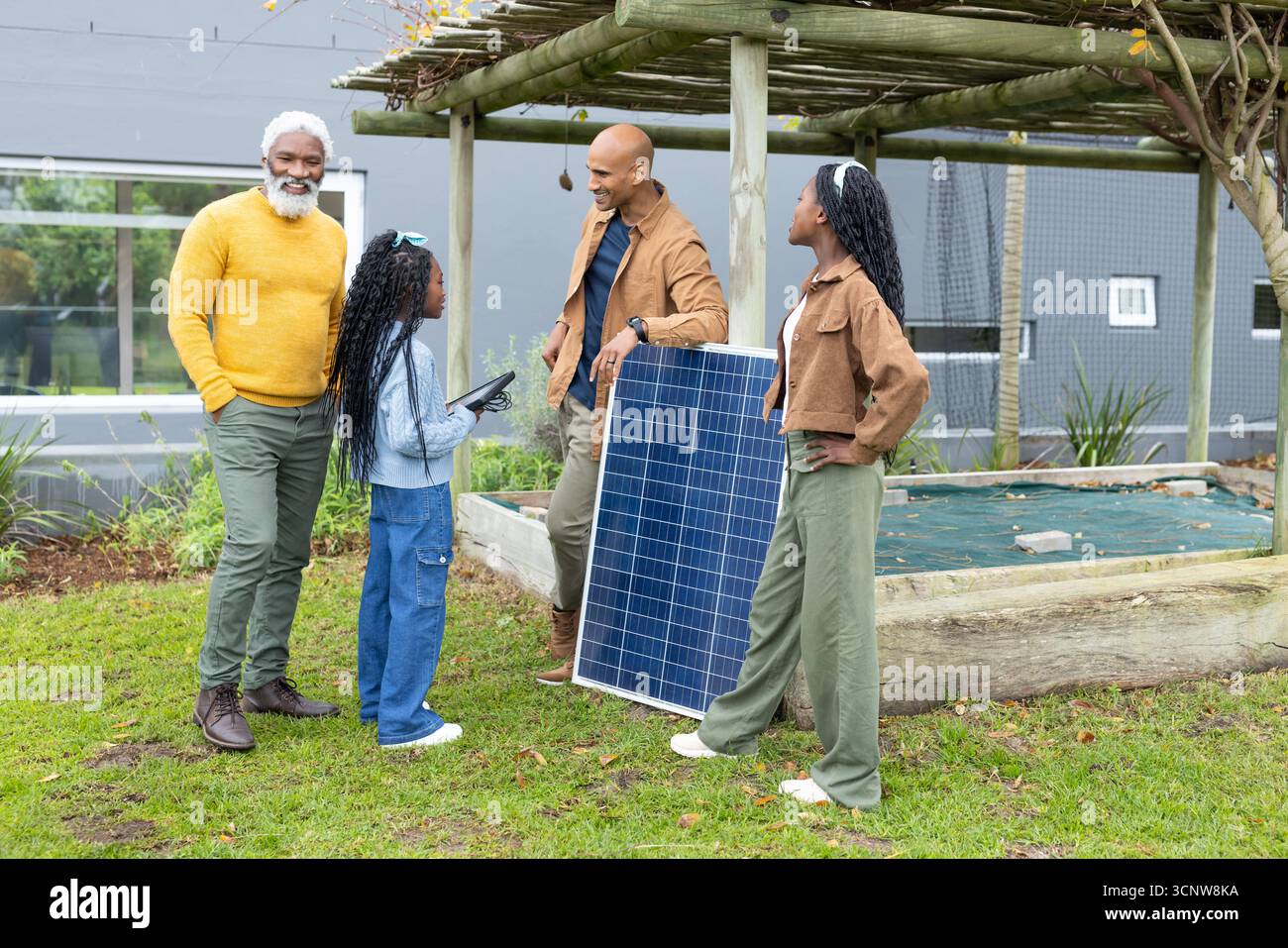 Diverse family gathering around solar panel under pergola on grassy lawn with tablet device Stock Photo