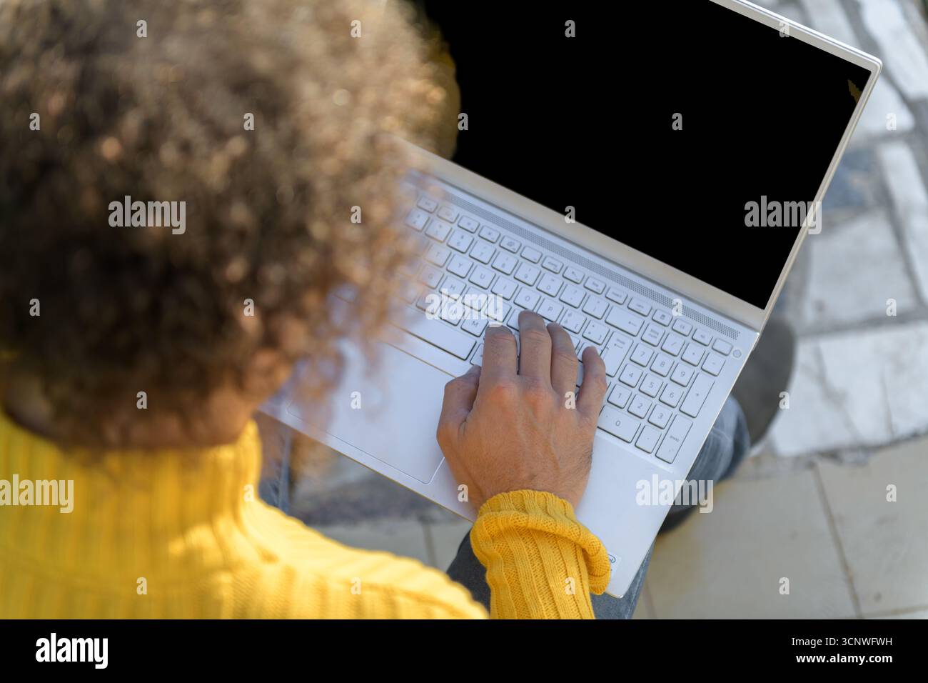 A man in a yellow sweater sits comfortably at home using a white laptop. The focus is on his hands typing on the keyboard, set against a relaxed, resi Stock Photo