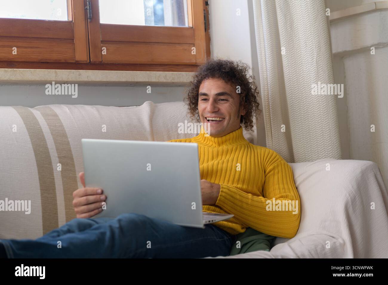 A man in a yellow sweater sits comfortably on a couch at home, engaged with his laptop. Natural light filters through a window, creating a cozy and in Stock Photo
