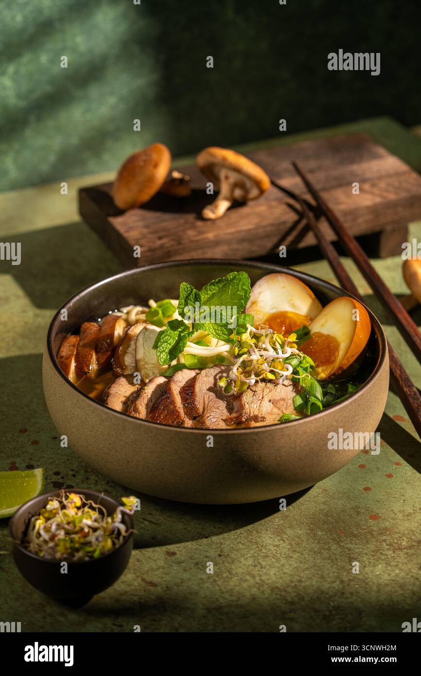 A delicious bowl of duck ramen features egg noodles, shiitake mushrooms, sliced duck, sprouts, and a boiled egg. Set on a rustic green background with Stock Photo