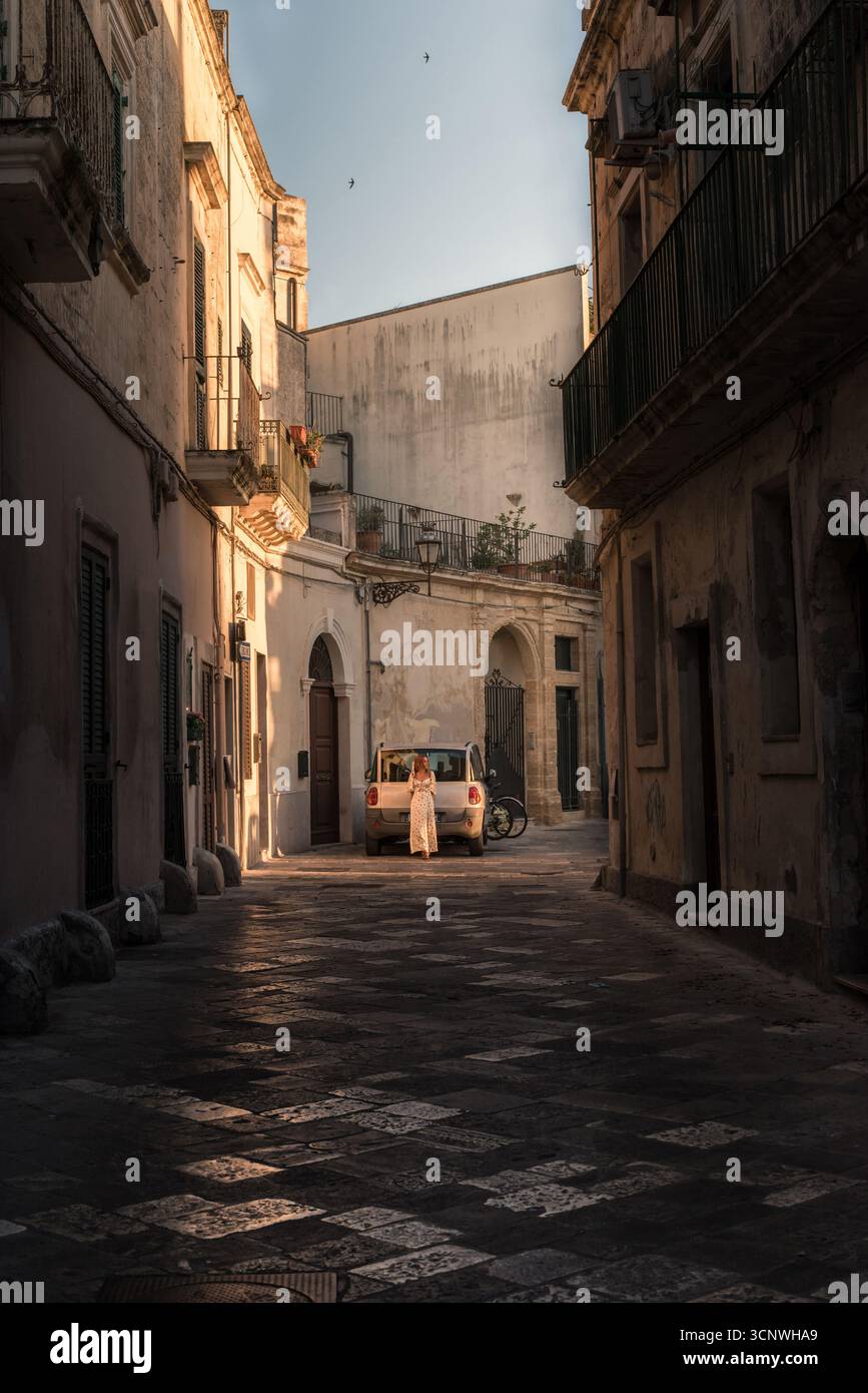 A sunlit alleyway in Lecce, Puglia, Italy, captures a serene scene with rustic architecture, soft shadows, and a parked car. A woman enhances the tran Stock Photo
