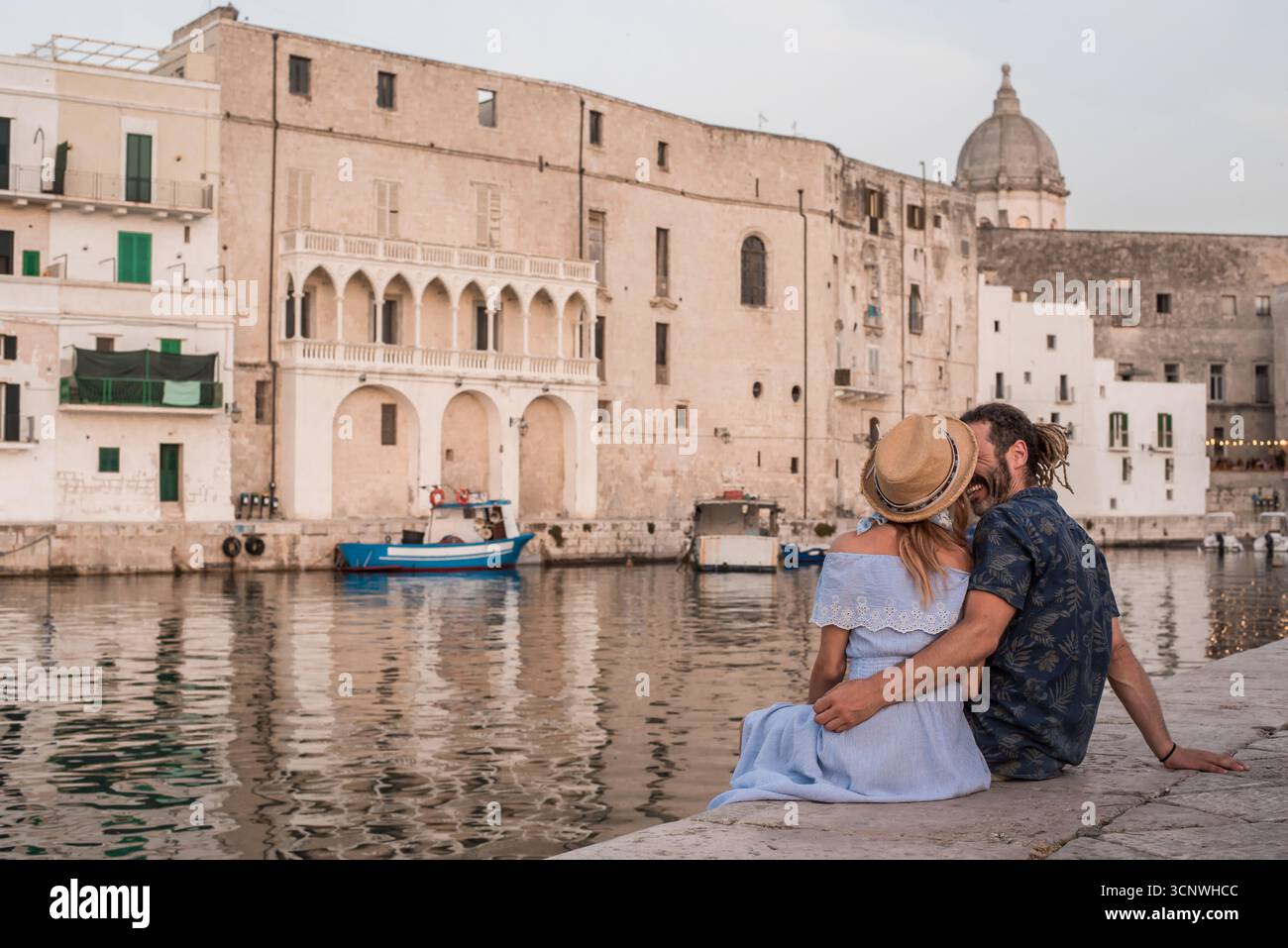 A couple sits by the tranquil waterfront in Monopoli, Puglia, Italy, embracing and admiring the historic architecture and serene water, enjoying a rom Stock Photo