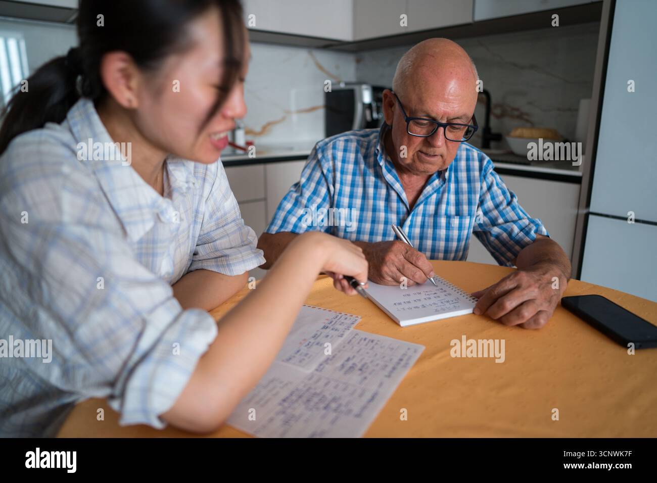 A senior man engaged in learning Mandarin, guided by an asian woman teacher, in a cozy kitchen setting. Both are focused on language notes and study m Stock Photo