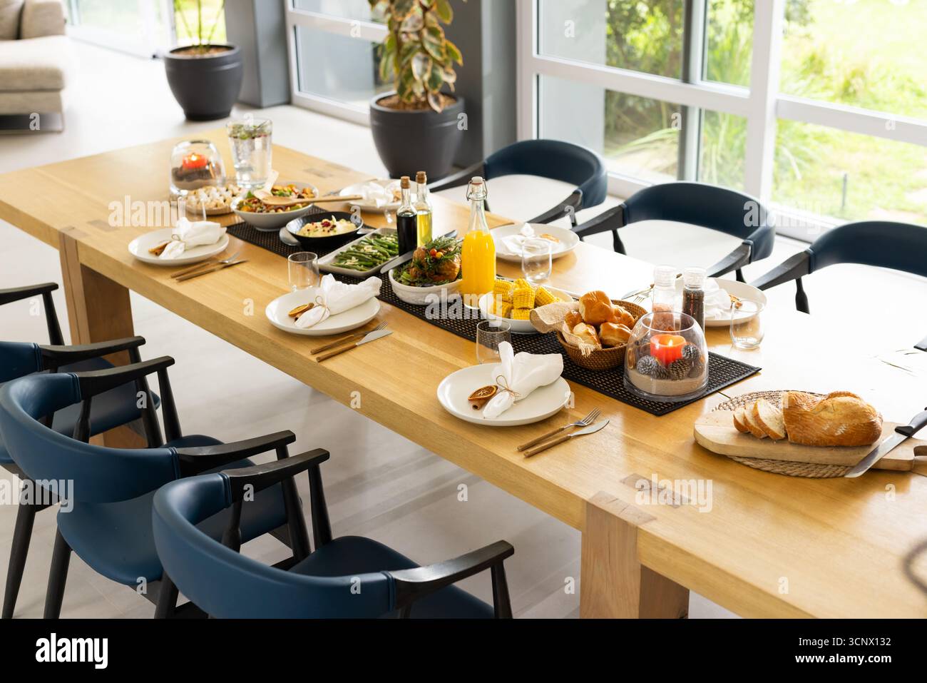 Wooden dining table hosting place settings, salads and breads, juice bottle, candles, navy chairs Stock Photo