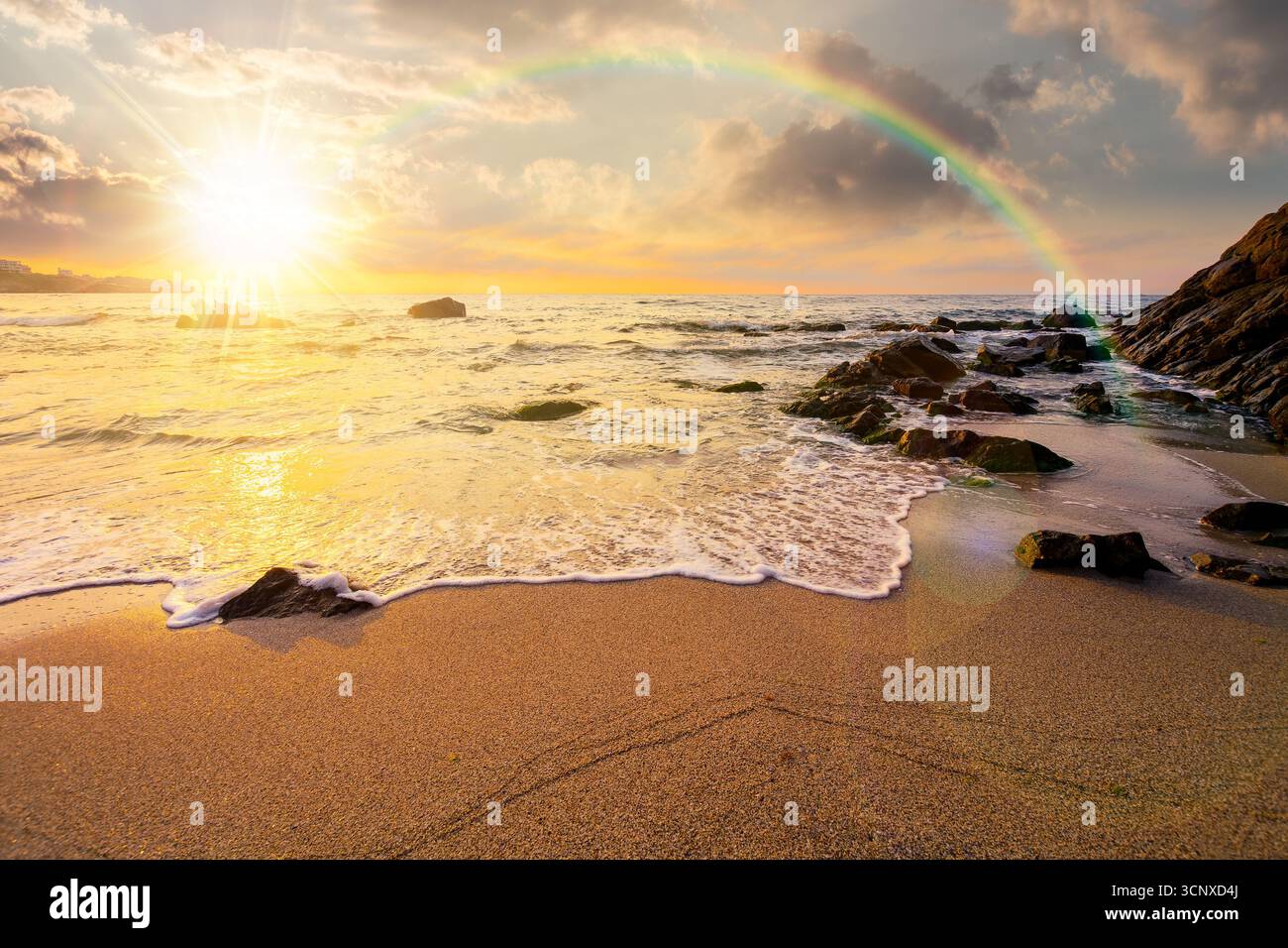 sea coast with rainbow over the boulders in the bay at sunset. sea tide with calm waves wash the golden sandy beach in evening light. beautiful autumn Stock Photo