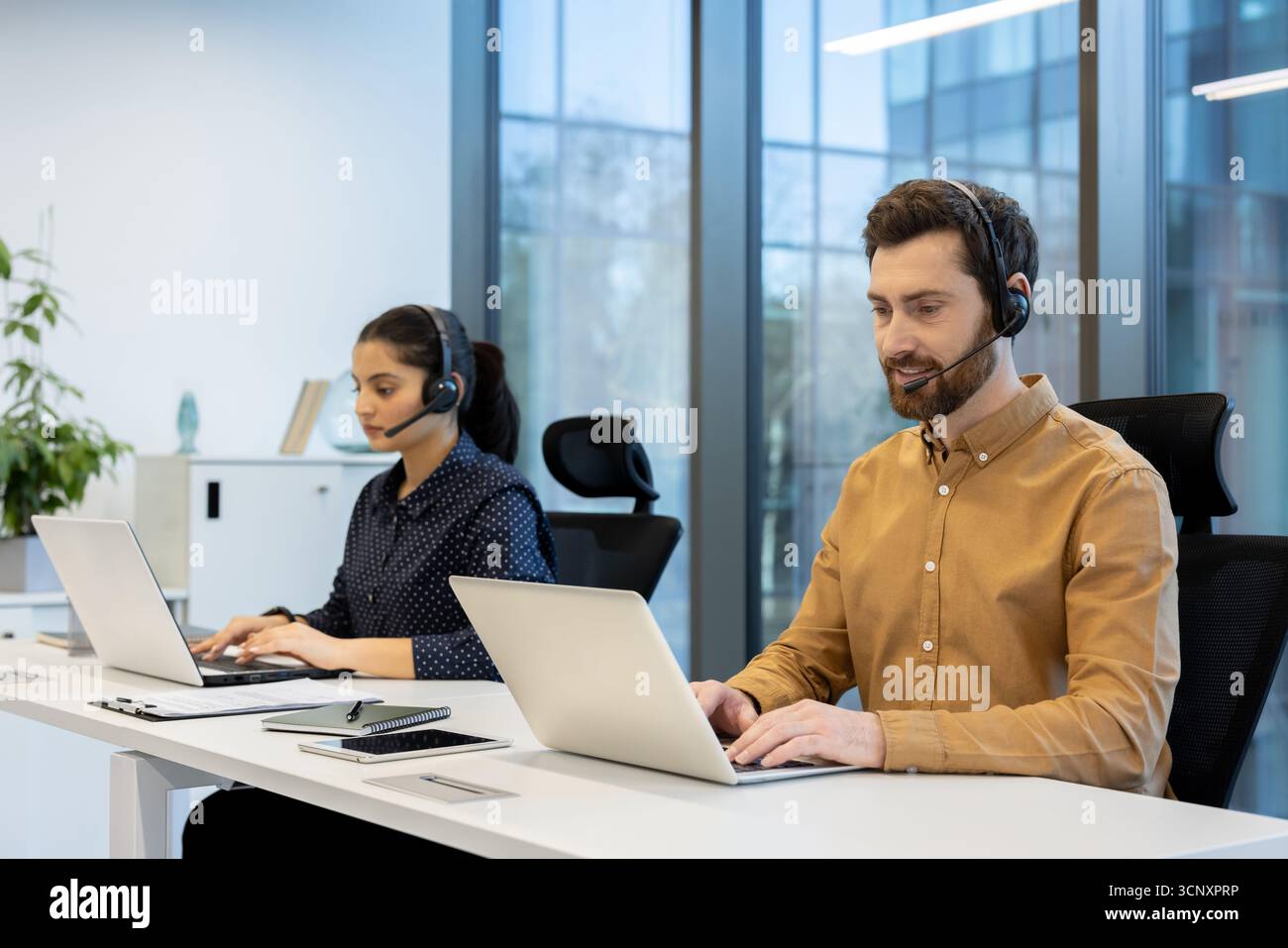 Two diverse call center operators wearing headsets and using laptops while working at desks in a bright, contemporary open-plan office, providing efficient telemarketing and online technical support Stock Photo