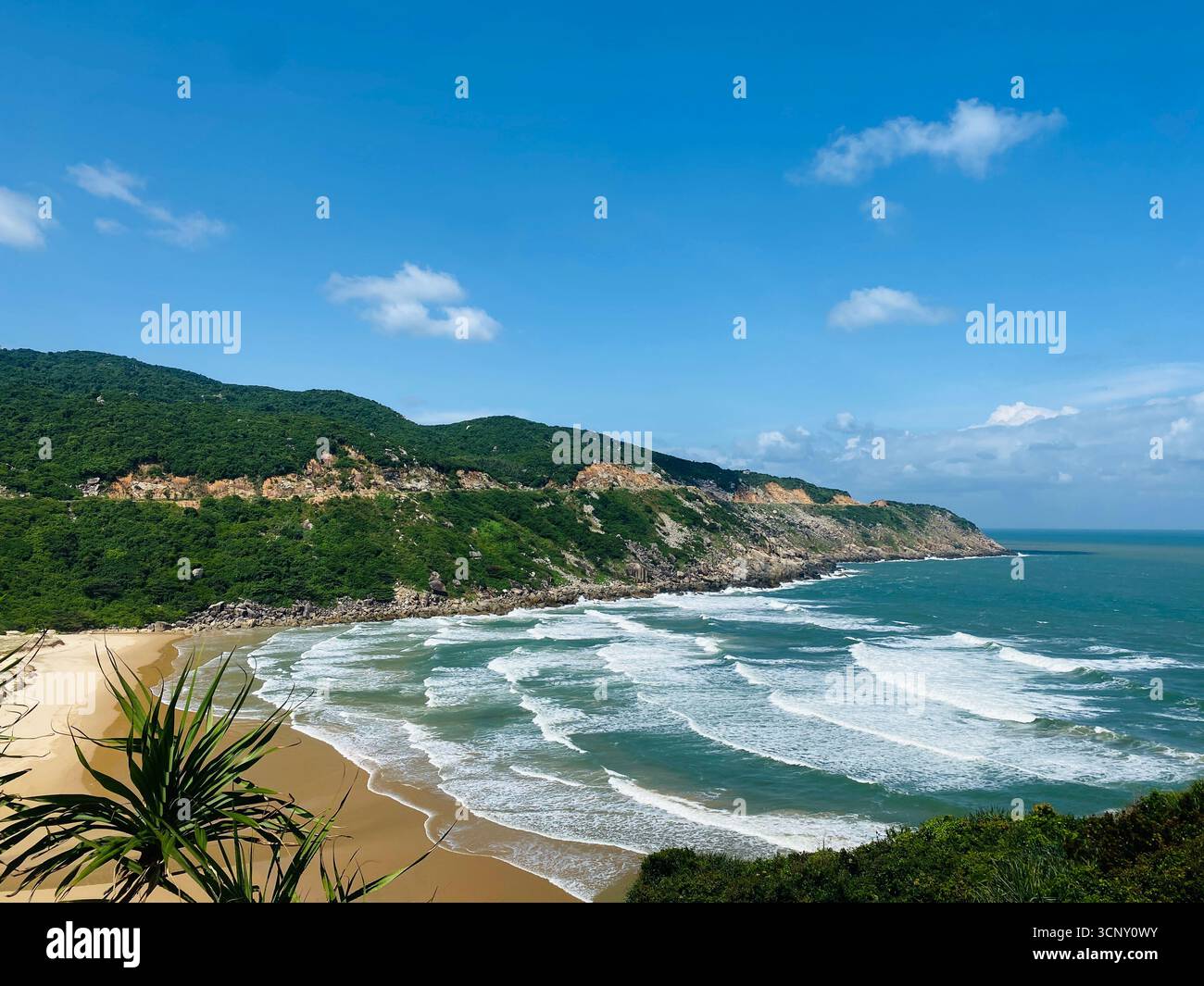 Scenic view of wild beach with turquoise waves, rocky cliffs, and lush green hills under blue sky in Vietnam. - Smartphone Captured Stock Image