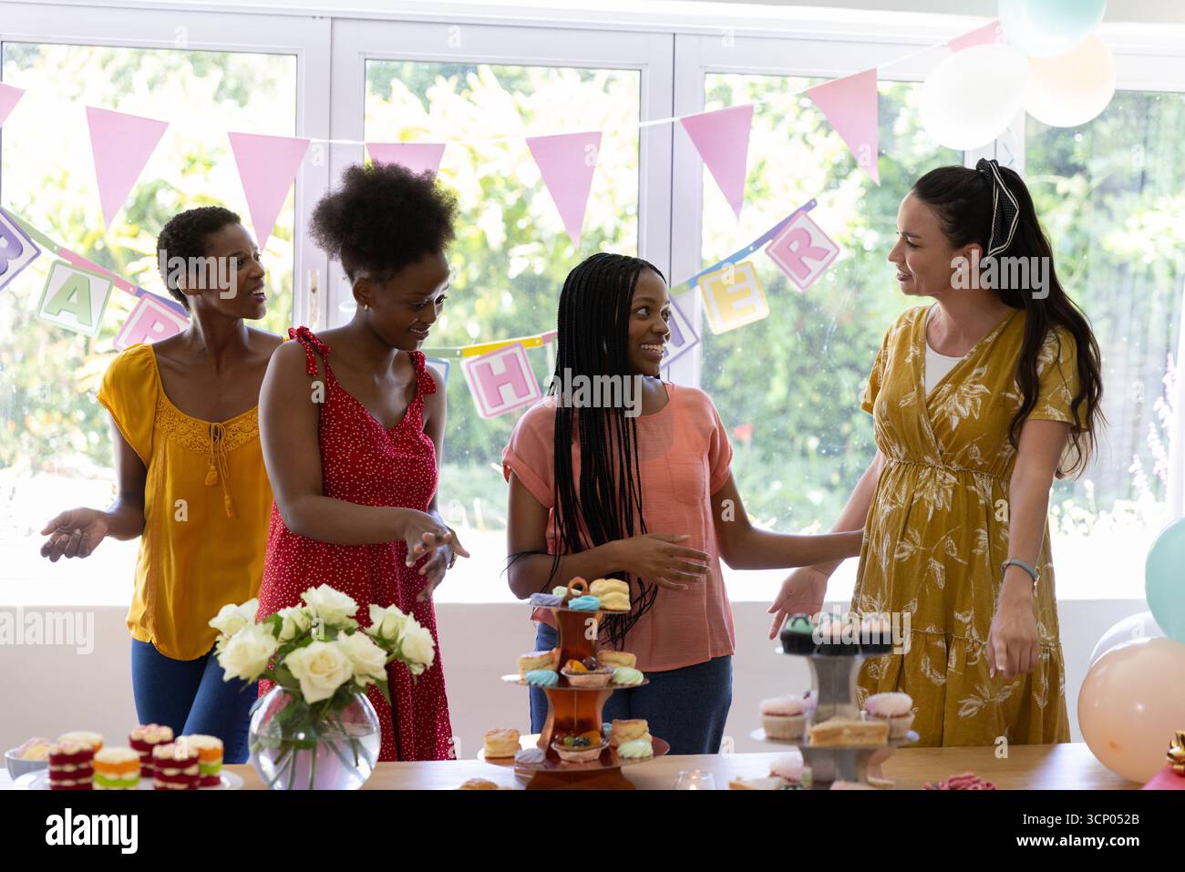 Diverse female friends gathering around table at home with tiered cupcakes and pink pennant banners Stock Photo