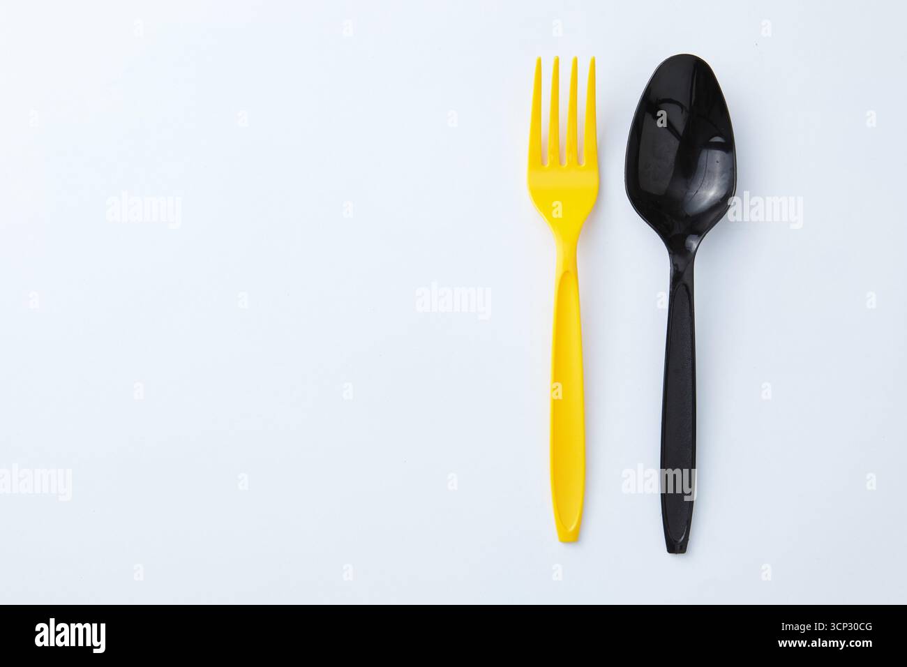 A yellow plastic fork next to a black plastic spoon on a white background Stock Photo