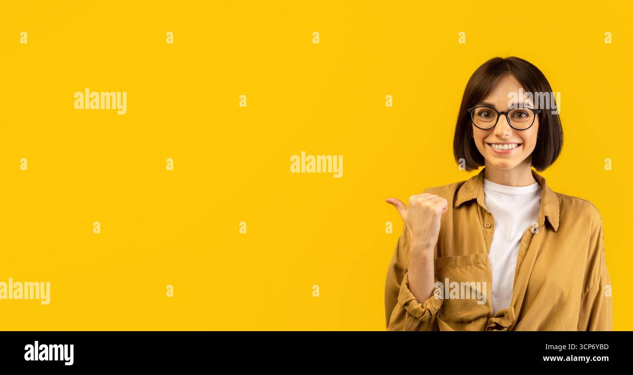 Check this offer. Positive young woman pointing thumb finger aside at free space, advertising your text or design Stock Photo