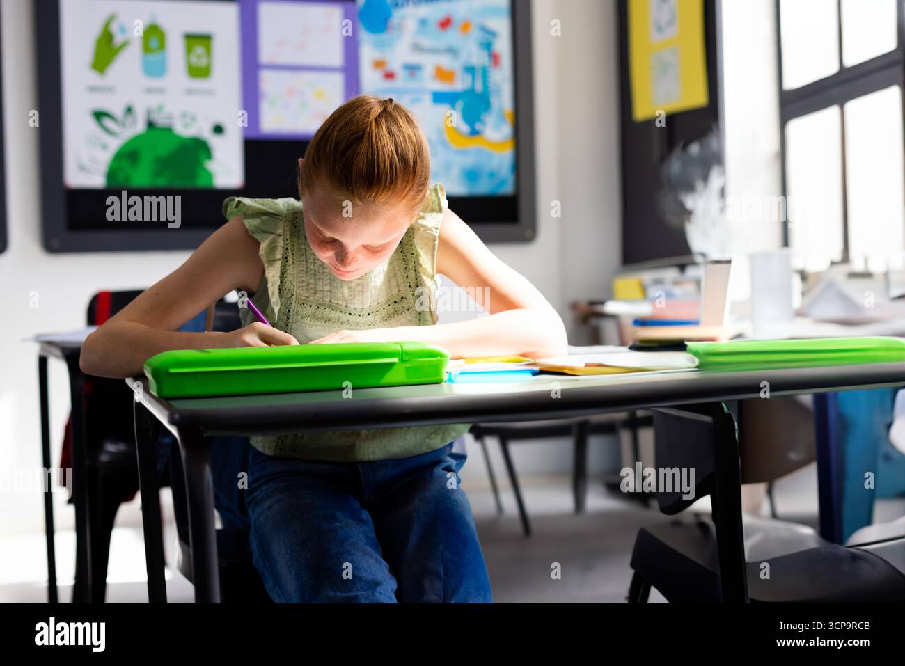 School-aged girl leaning forward at desk in classroom, writing in notebook with green pencil case Stock Photo