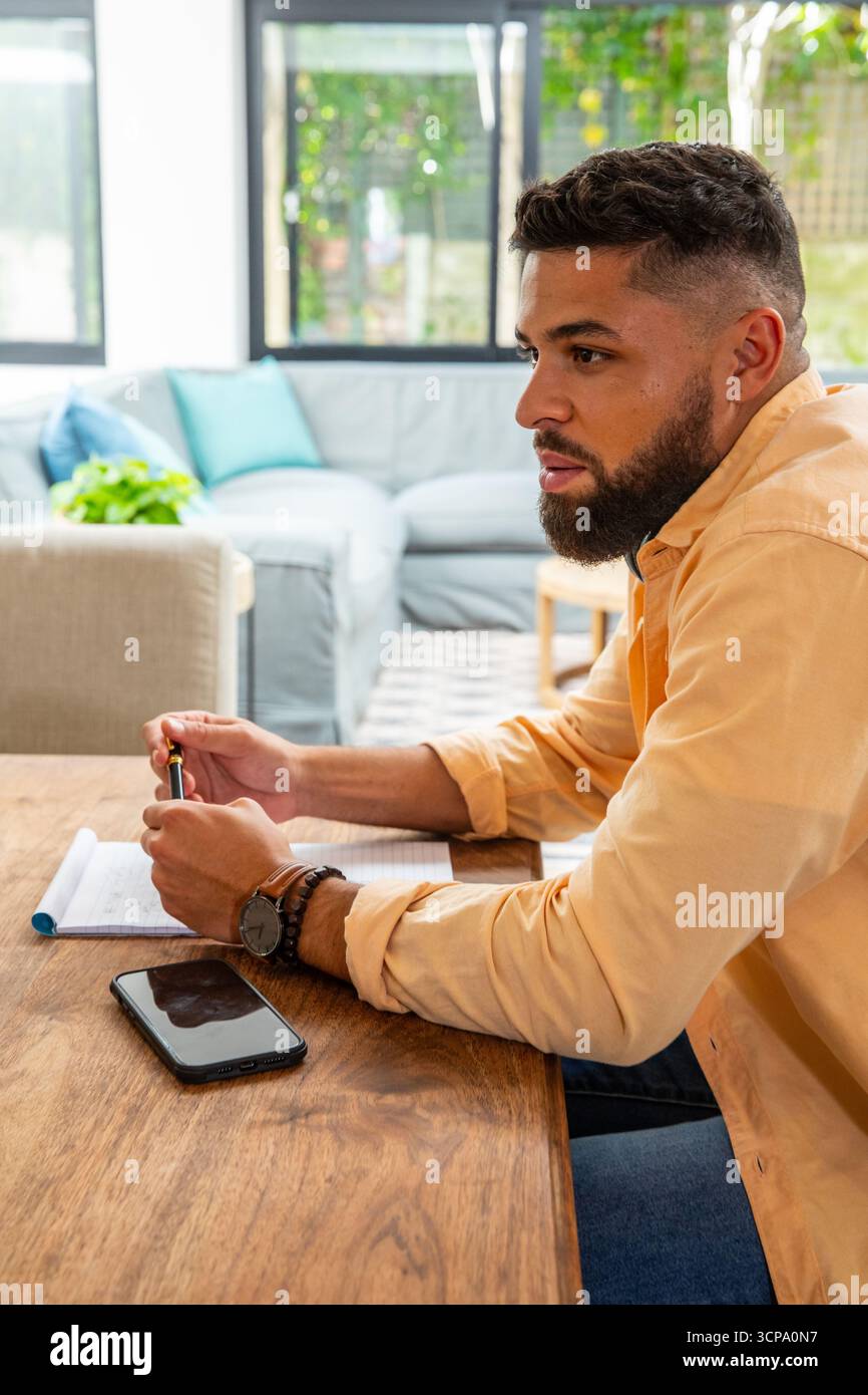 Man scribbling in notebook while leaning on wooden table at home with smartphone Stock Photo