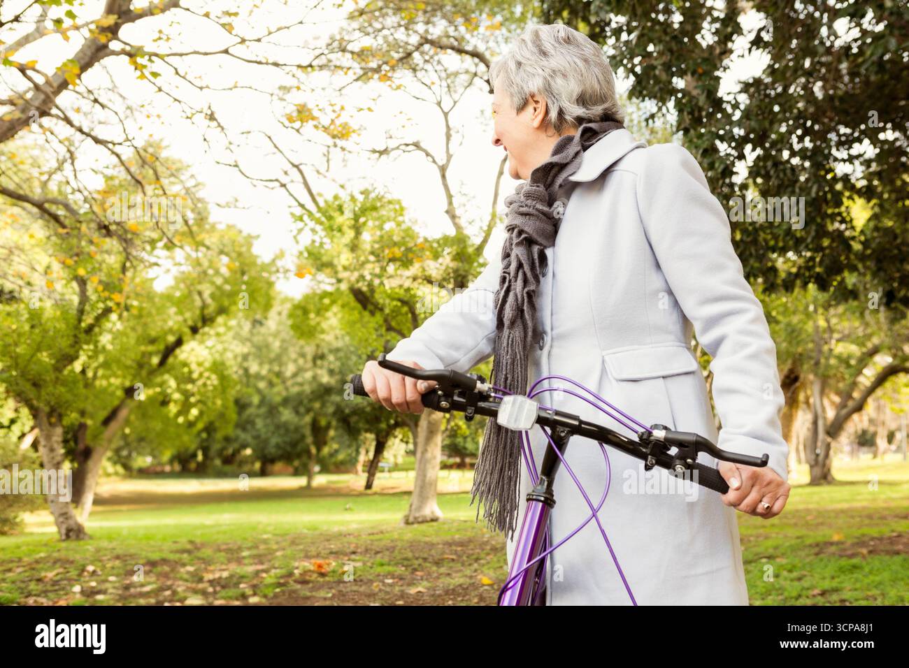 Senior woman holding bicycle and wearing long coat with scarf in leafy autumn park, copy space Stock Photo