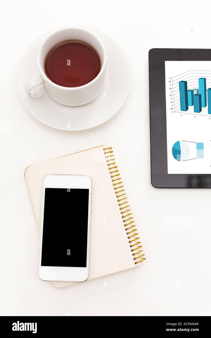Busy workspace with coffee, smartphone, notebook, and tablet displaying data graphs Stock Photo