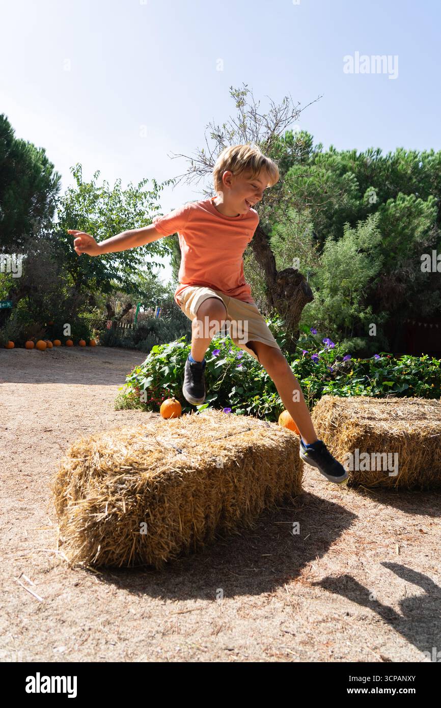Blond boy actively jumping over a hay bale in an outdoor pumpkin patch setting Stock Photo