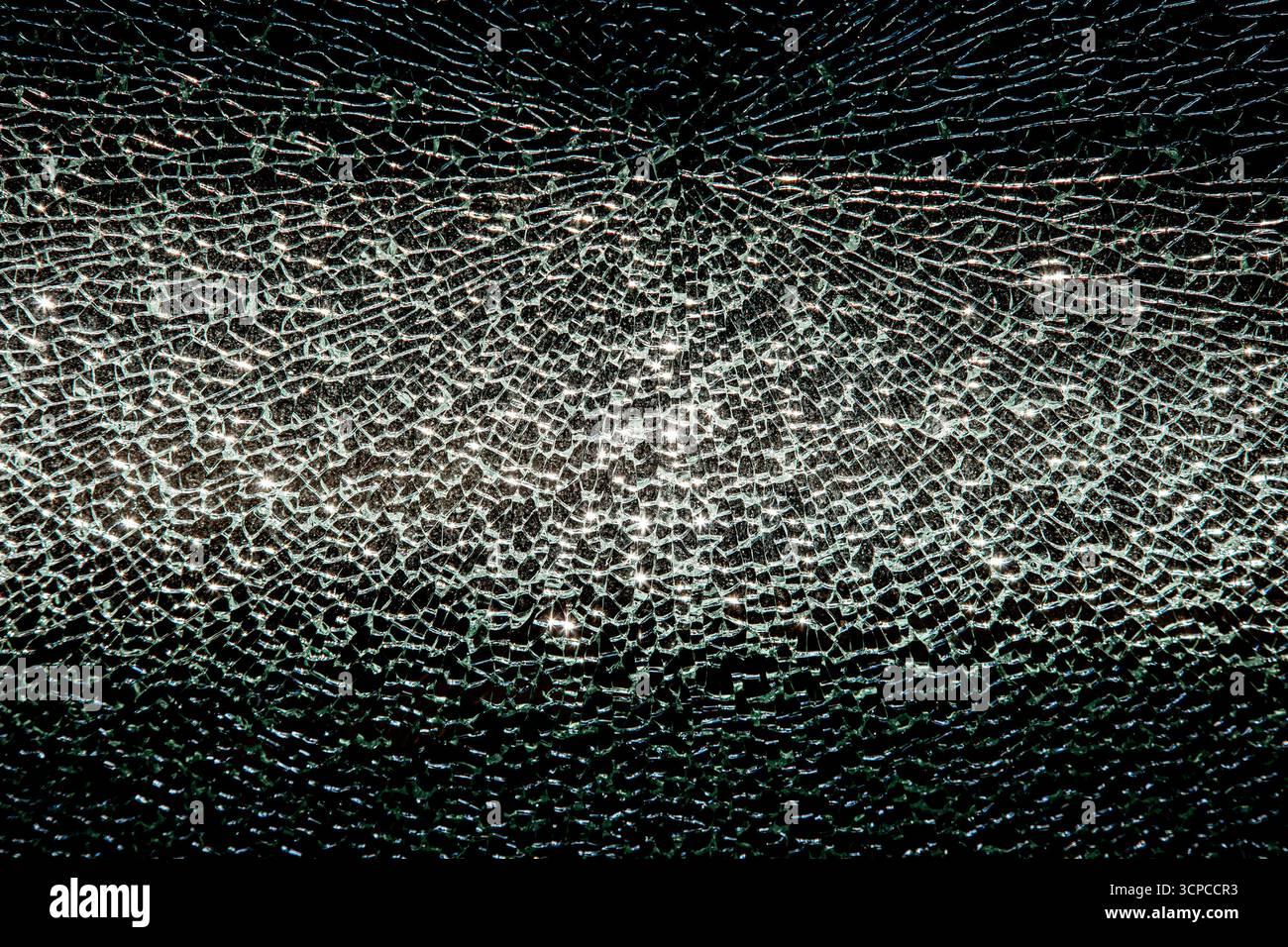 Close-up of a broken glass window showing a complex web of cracks radiating from a central impact point. The jagged shards reflect light in greenish-w Stock Photo