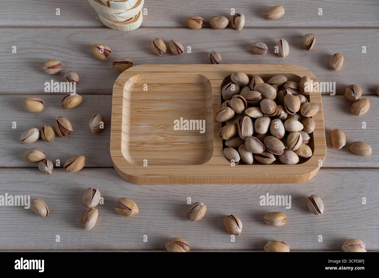 A top-down view of rustic shelled pistachios on a wooden tray, scattered on a light wooden surface, highlighting natural textures and earthy tones Stock Photo