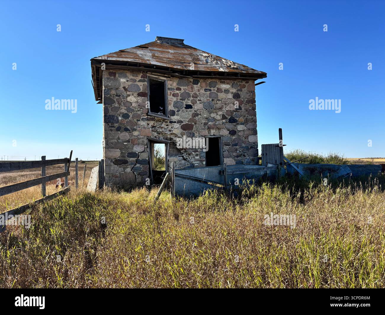 Close-up view of an abandoned historic stone farmhouse surrounded by tall prairie grass and rustic wooden fencing under a bright blue sky. - Smartphone Captured Stock Image