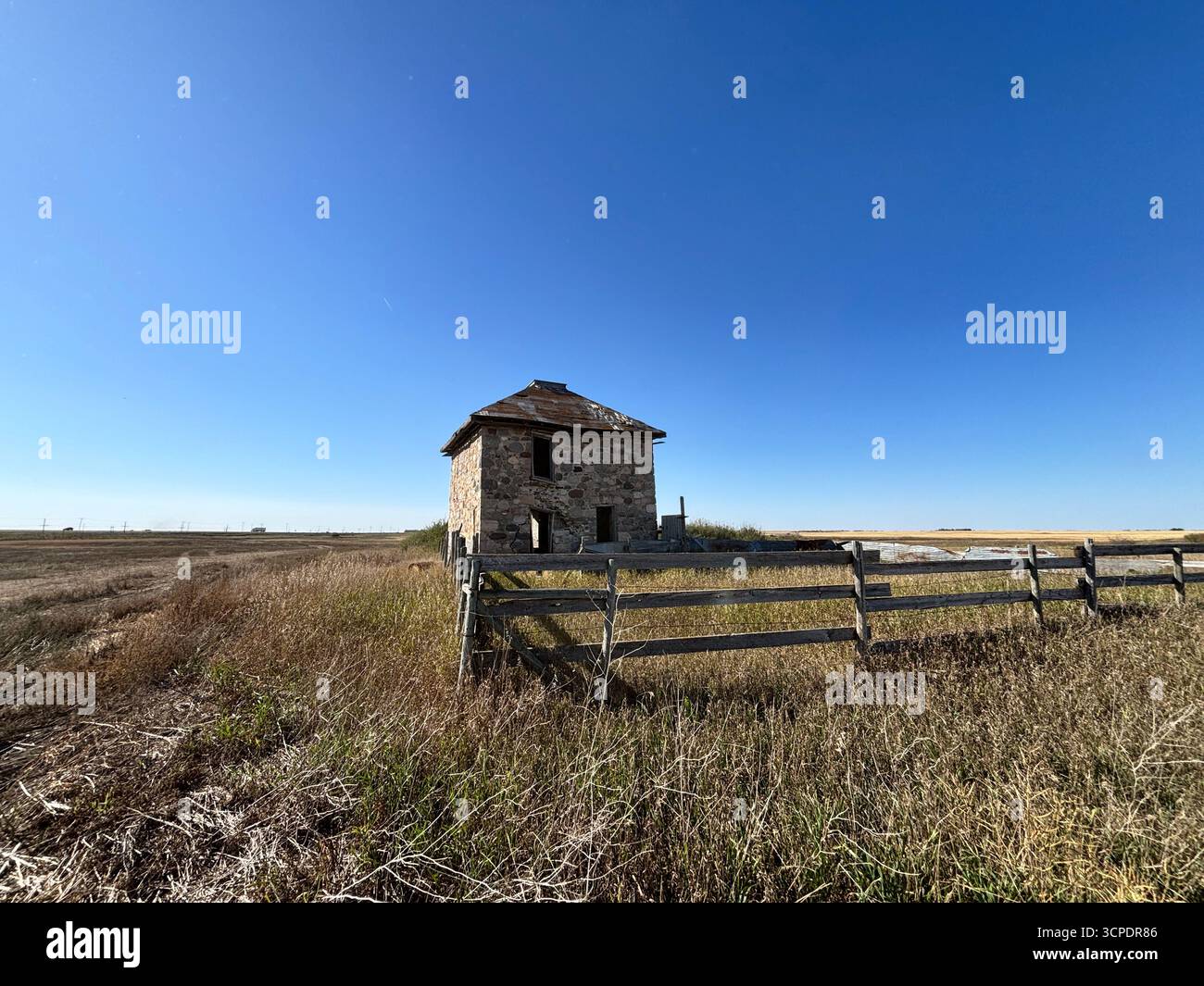 View of an abandoned stone farmhouse with weathered wooden fencing and overgrown prairie grass under a vast blue sky in rural Saskatchewan, Canada. - Smartphone Captured Stock Image