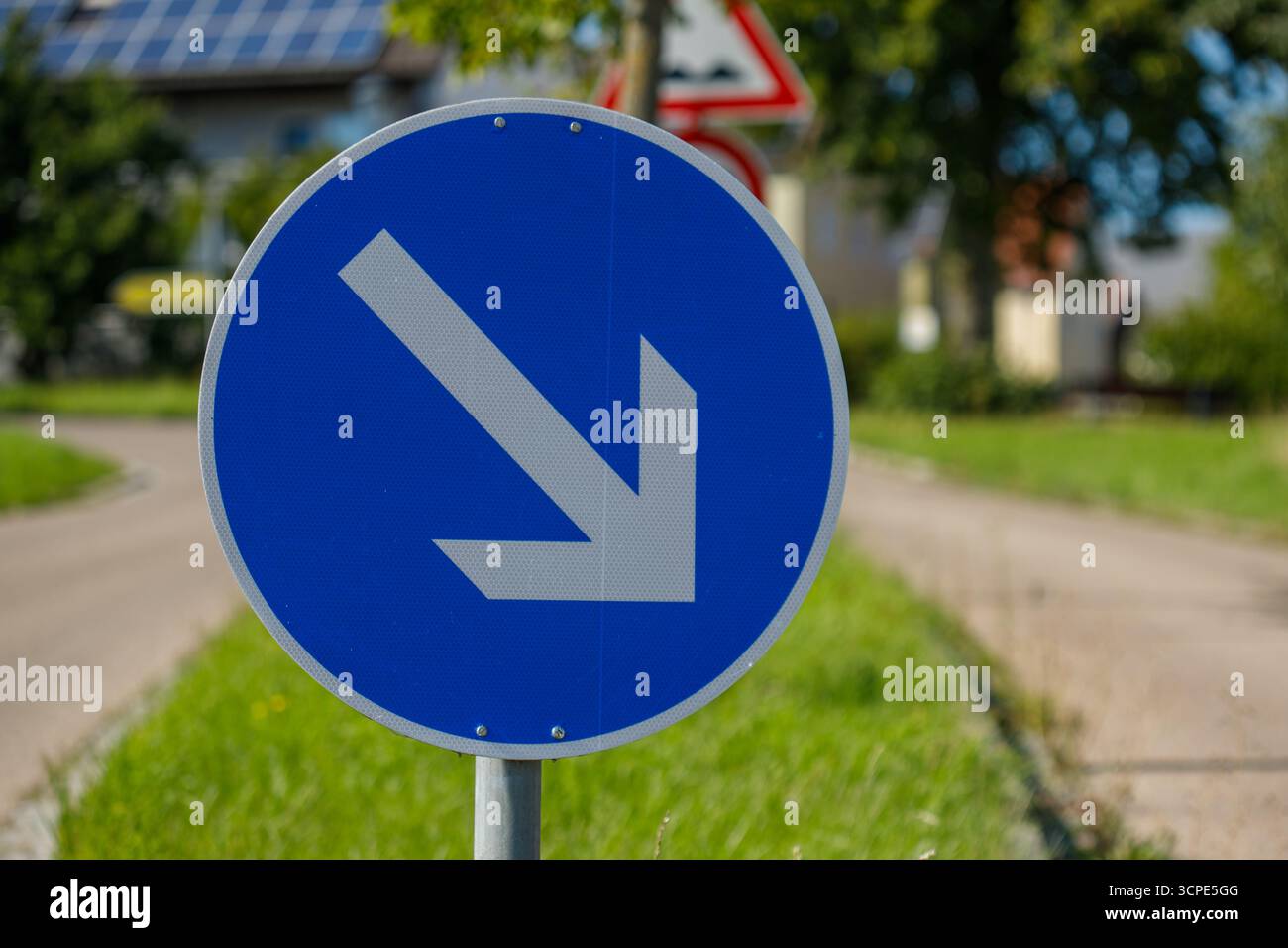 A close-up shows a blue and grey road sign with a directional arrow pointing downwards and to the left, against a blurred background. Stock Photo