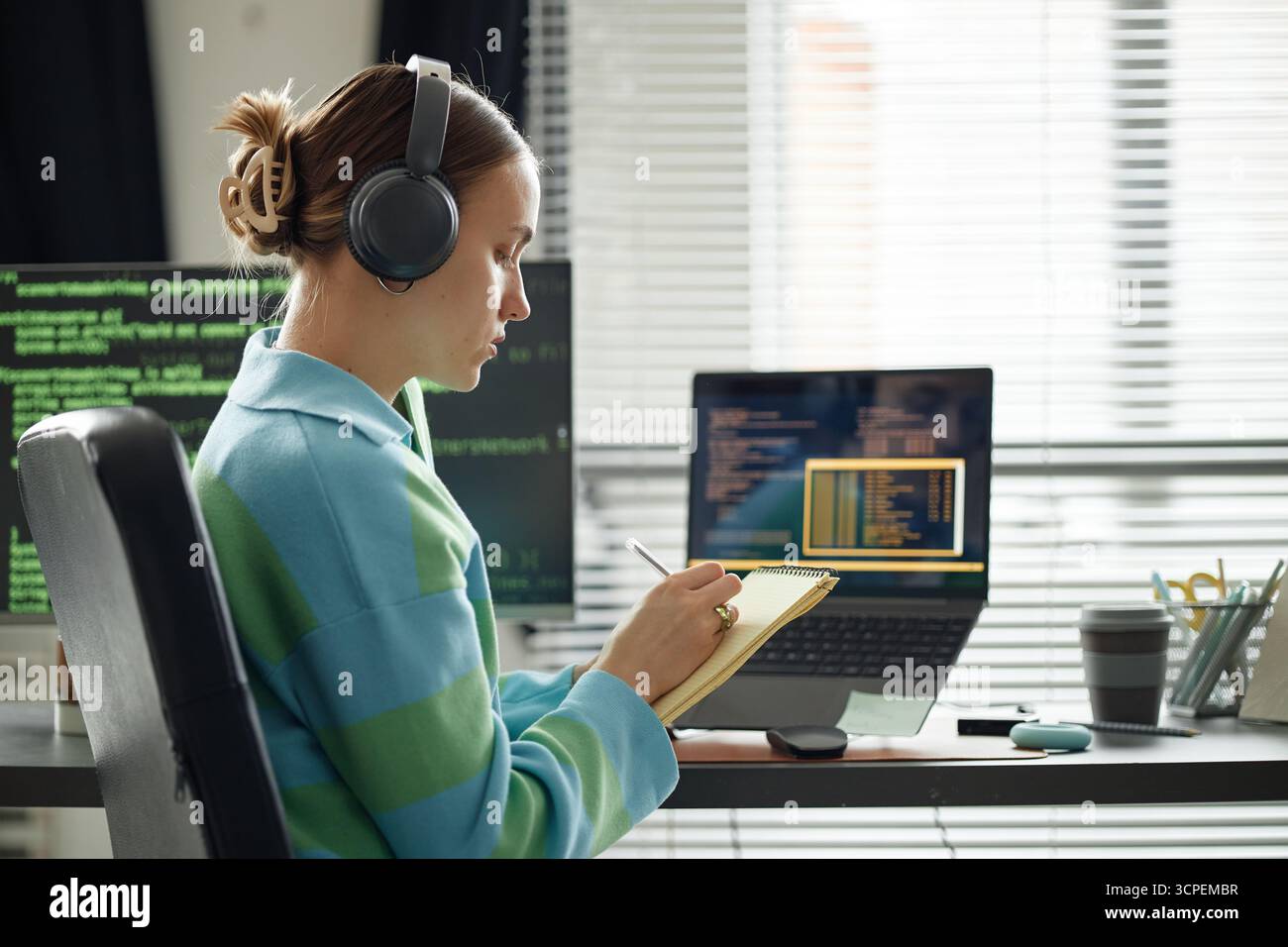 Young Woman Wearing Headphones Writing Notes While Coding at Desk Stock Photo