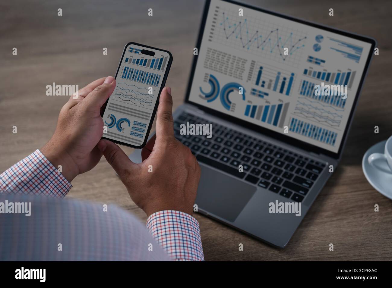 hands holding smartphone with dashboards, laptop screen showing charts and graphs in a modern workspace; data, metrics, and reports for business perfo Stock Photo