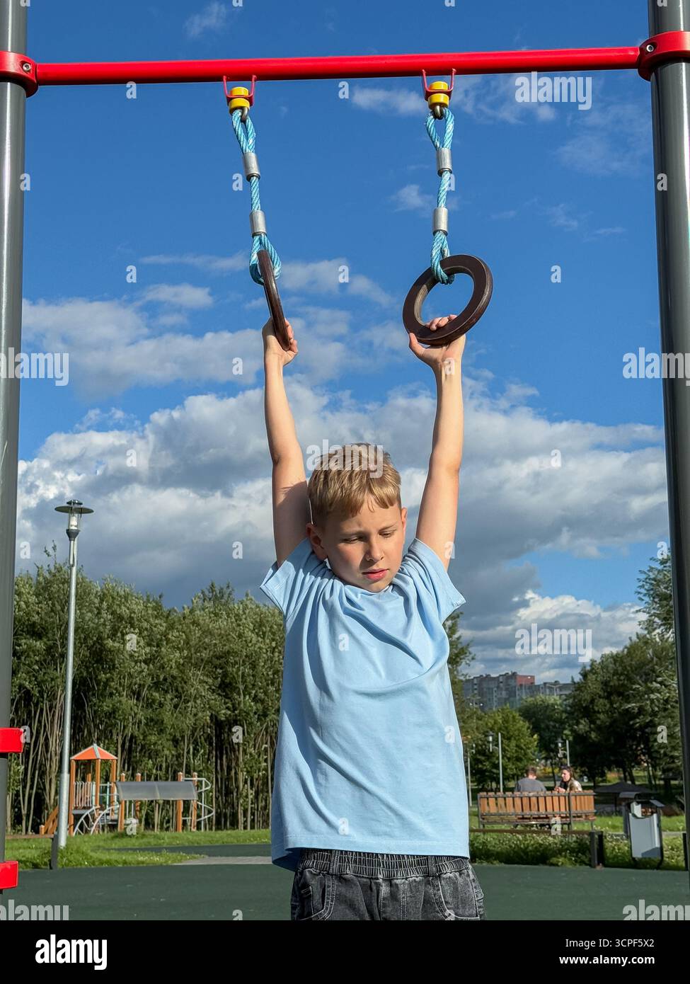 Teen boy exercising on gymnastic rings at an outdoor sports playground during a bright summer day, fitness and athletic activity concept - Smartphone Captured Stock Image