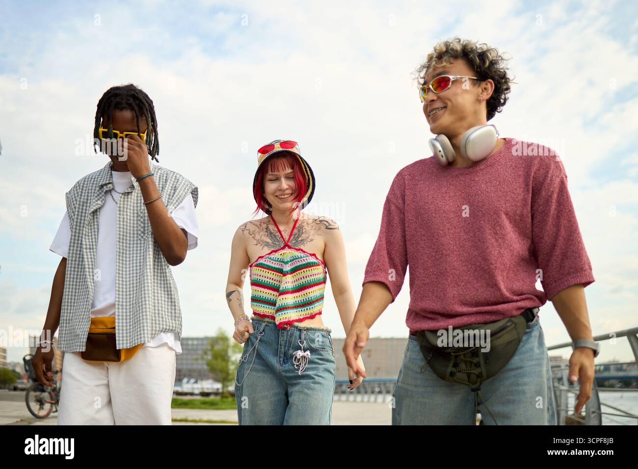 Group of three young people, multiethnic men and Caucasian woman, walking outdoors together, smiling and interacting, wearing sunglasses and headphones, enjoying urban environment Stock Photo