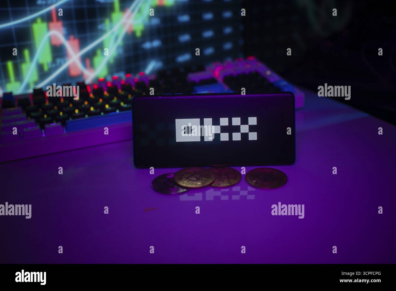 OKX logo displayed amidst purple hue with coins and trading graphs visible behind Stock Photo