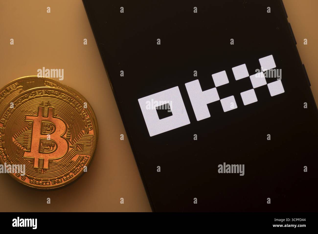 Bitcoin coin next to a smartphone featuring the OKX logo on a dark background Stock Photo