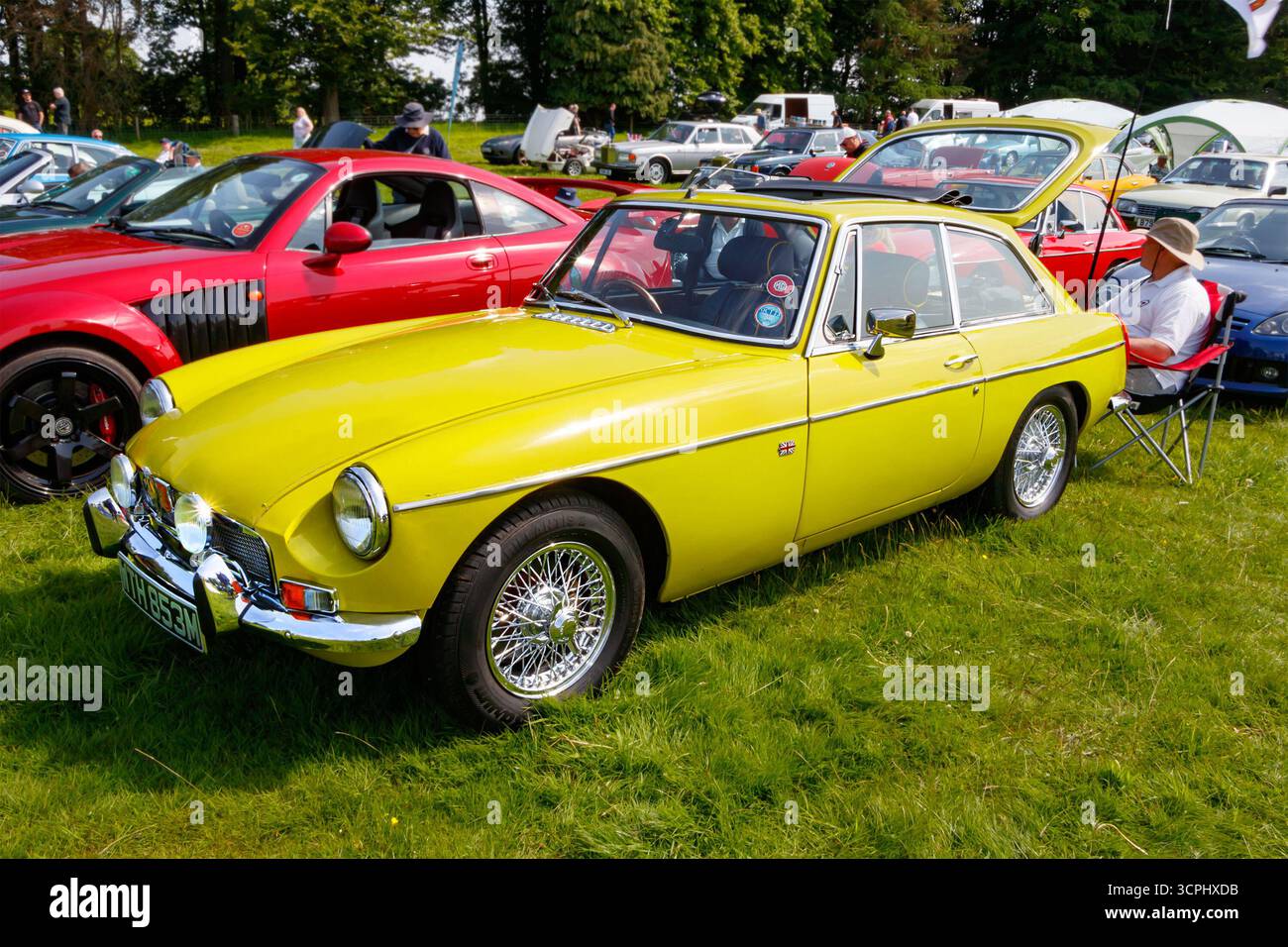 Vintage yellow MGB sports car displayed on a grassy field at a classic car show with other vehicles in the background Stock Photo