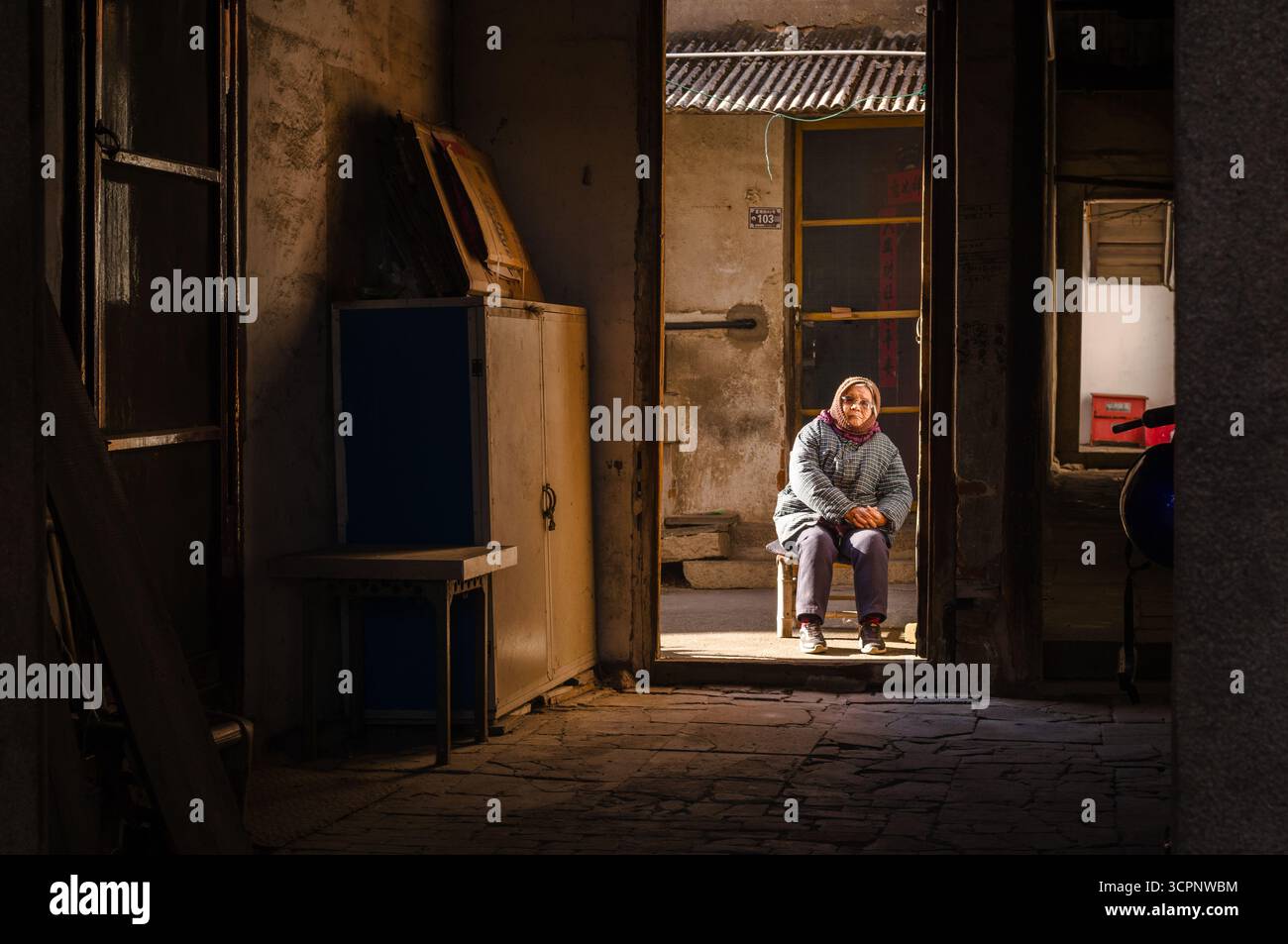 An elderly woman sits in the sunlit doorway of a weathered, traditional building, with light streaming into the dim, rustic interior. Stock Photo