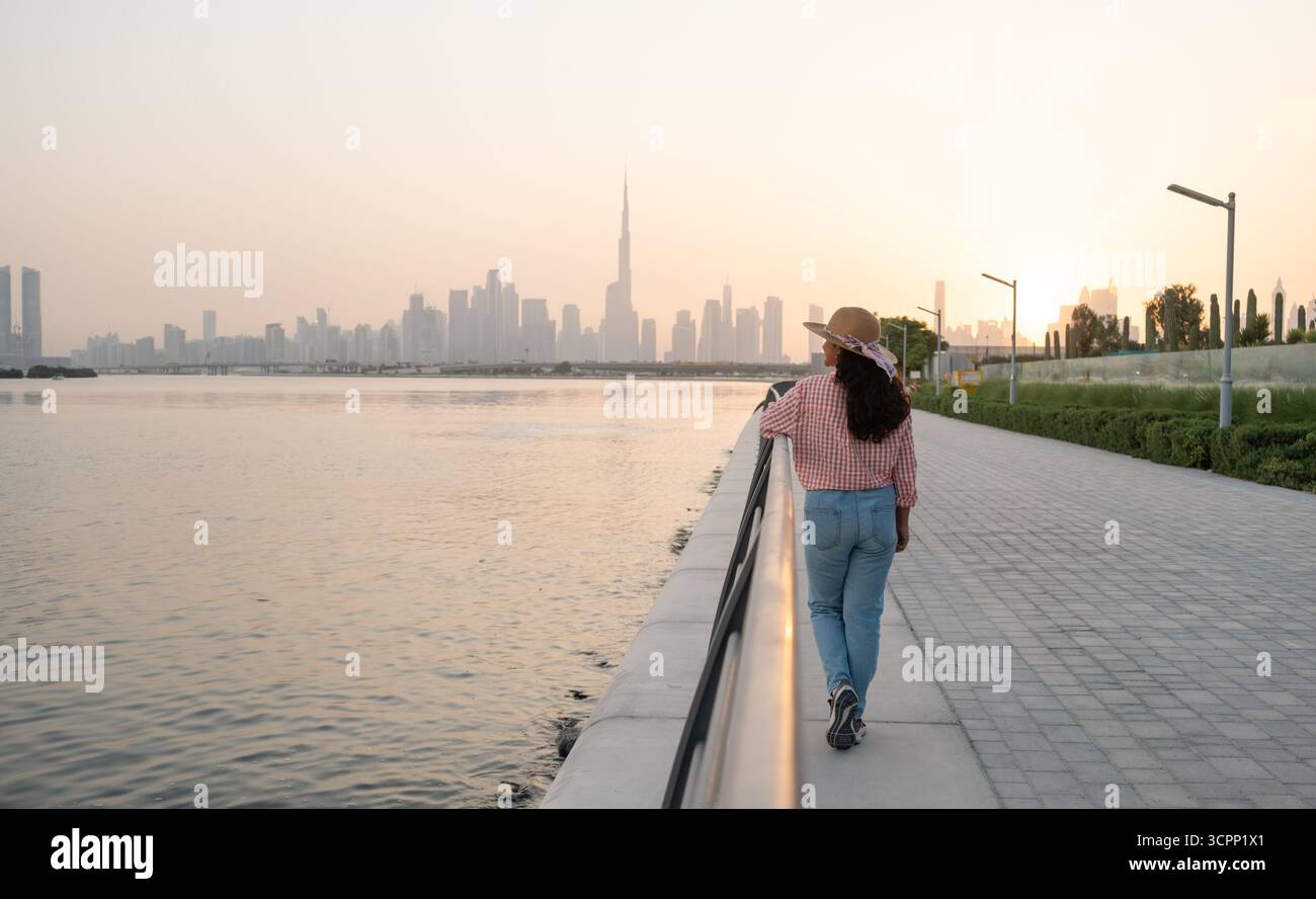 Dubai skyline silhouette at sunset with a woman wearing a sunhat and shirt standing at the waterfront promenade Stock Photo