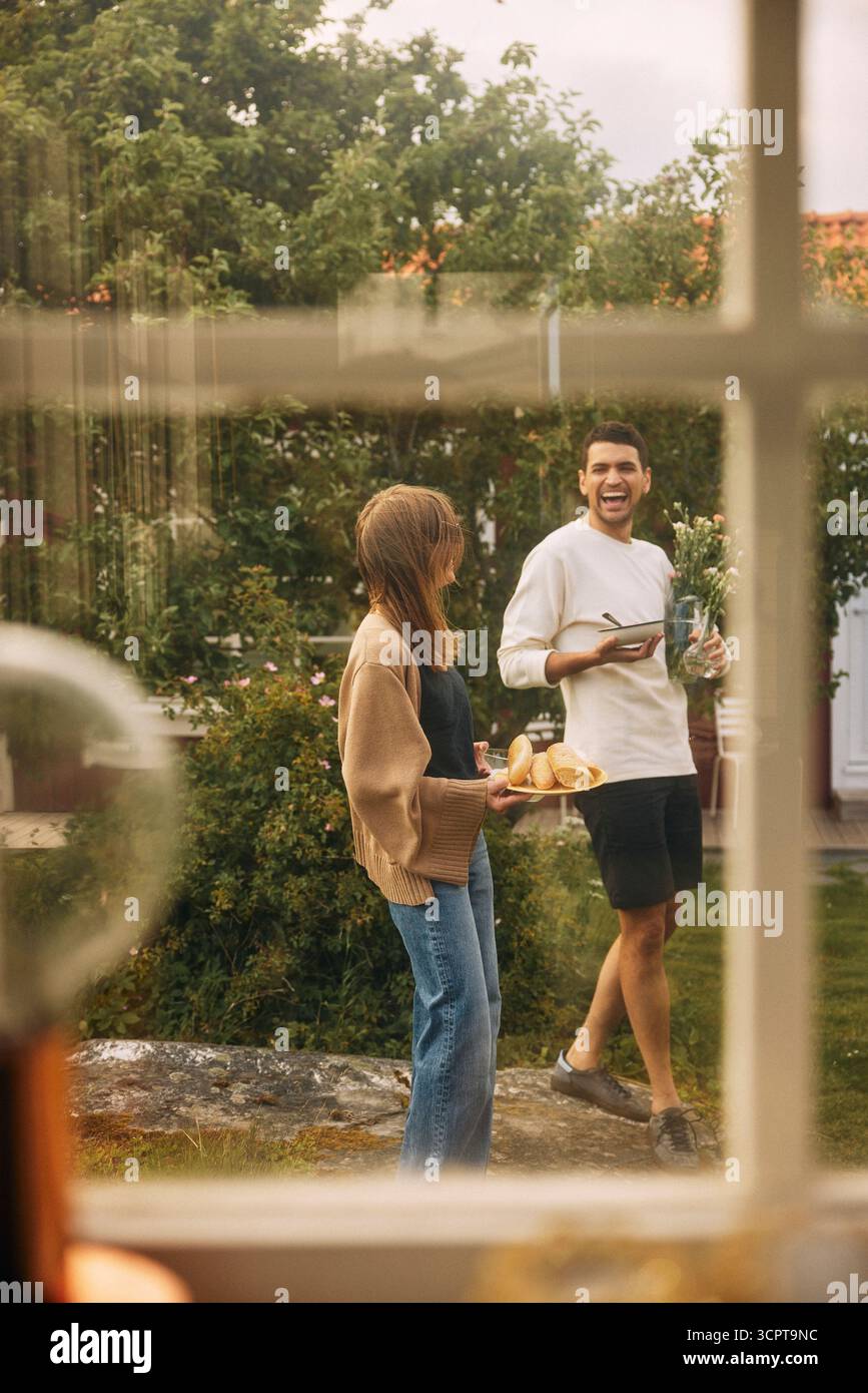 Carefree man enjoying with female friend holding plate of bread while walking in back yard seen from window Stock Photo