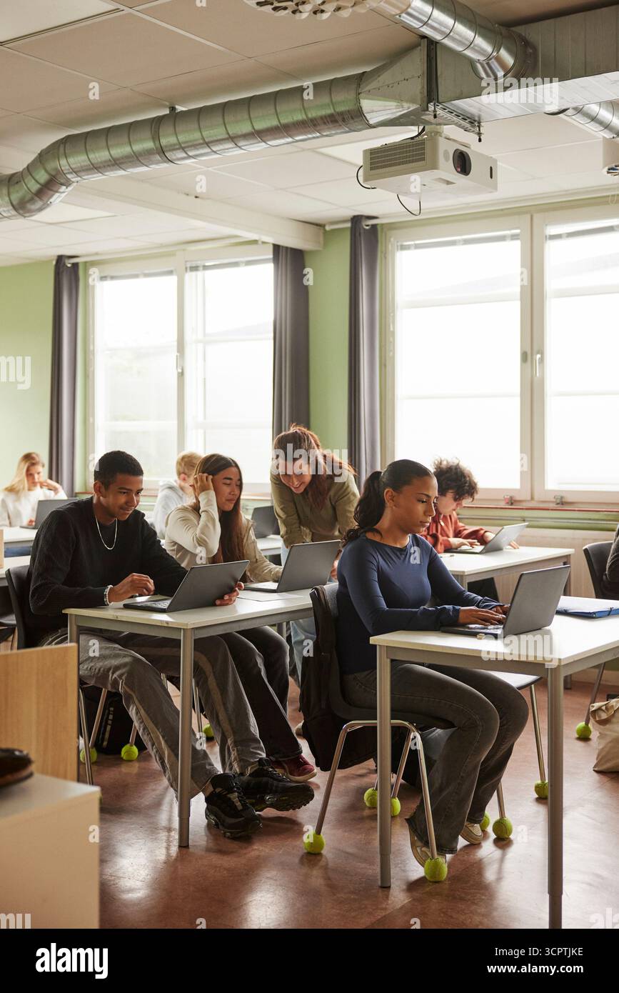 Female teacher assisting students doing e-learning in classroom at junior high school Stock Photo