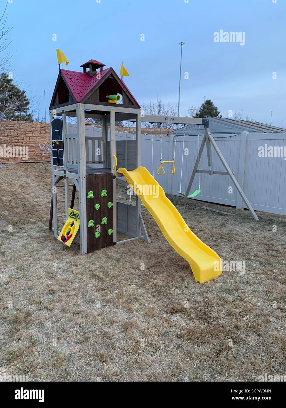 A wooden play structure stands in a backyard during the early spring, The play set includes a rock wall, slide, swings, and a clubhouse. - Smartphone Captured Stock Image
