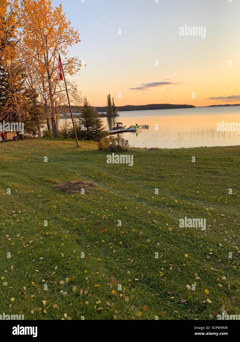 A tranquil lake sunset illuminates fall foliage and a dock with small boats. A Canadian flag stands tall in the warm, golden light.peaceful - Smartphone Captured Stock Image