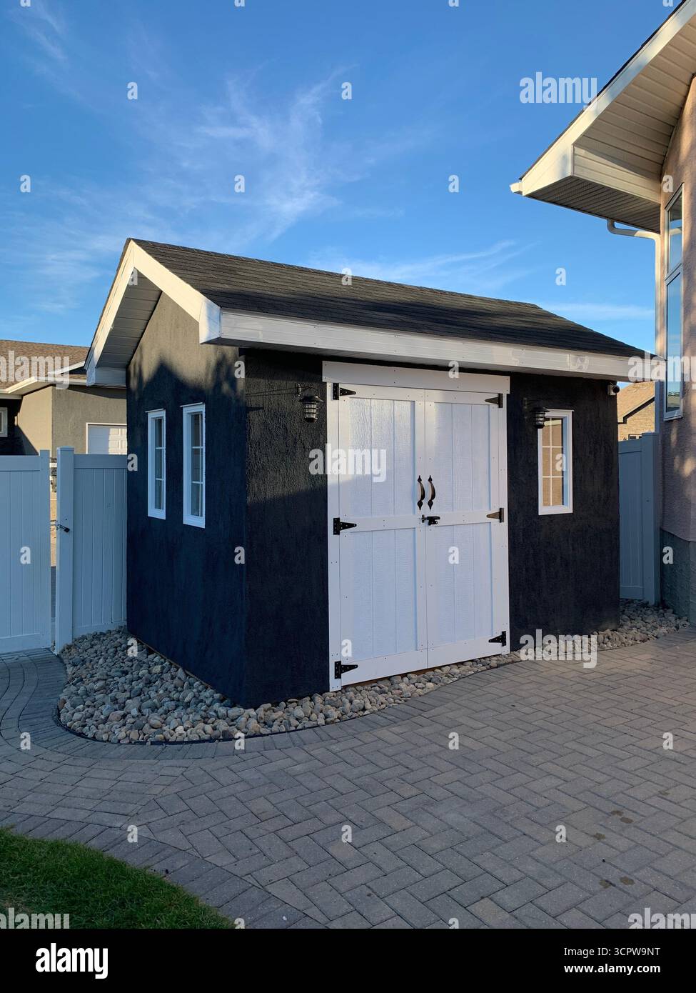 Modern garden shed with white double doors and windows, surrounded by gravel landscaping on a sunny patio near home exterior. - Smartphone Captured Stock Image