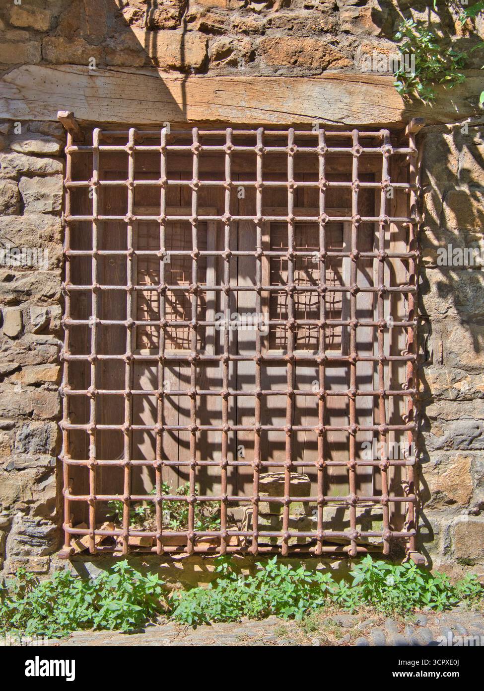 Rustic Window with Wrought Iron Grate - Close-up of an old wooden window with a rusty square wrought iron security grate set into a rough, historic st Stock Photo
